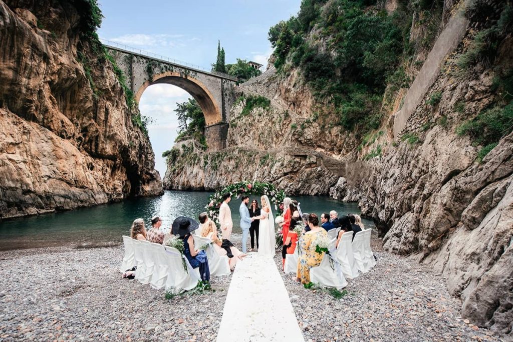 Italian Wedding Photographer captures a wedding group on a Positano beach with a stone bridge, showcasing the dramatic coastal scenery