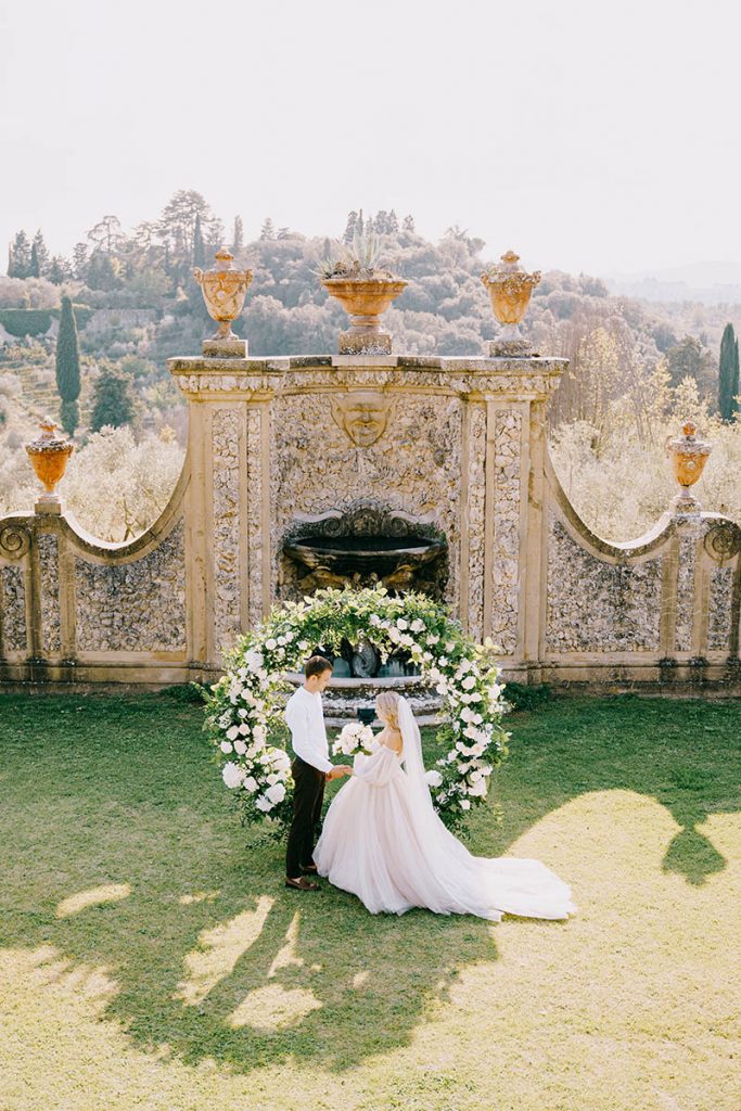 Bride and groom exchanging vows in front of ornate fountain and floral arch, Umbria wedding photographer style