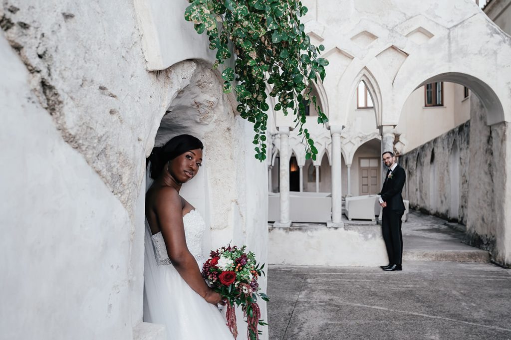 Bride holding a bouquet in a historic courtyard with an arched doorway and groom in the background