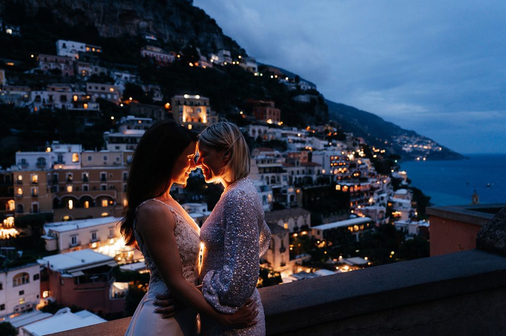 Romantic couple embracing with the twinkling lights of Positano at night in the background.