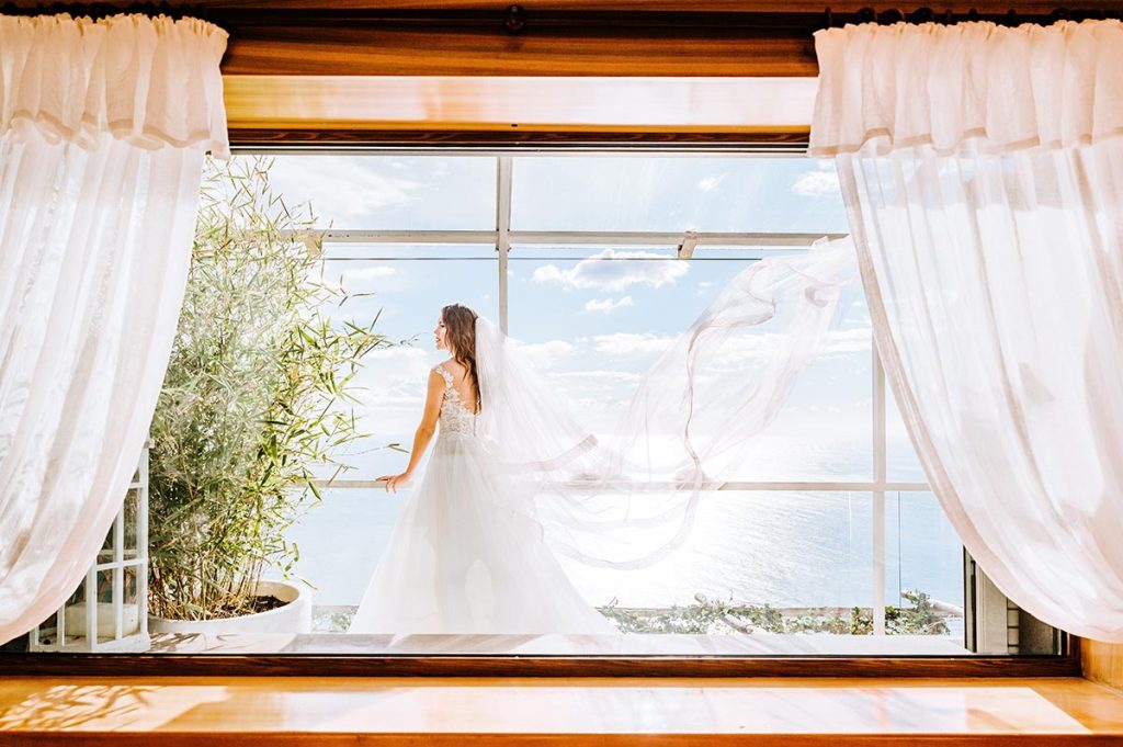 Bride seen through a window with flowing curtains, highlighting the picturesque settings curated by a Capri wedding planner
