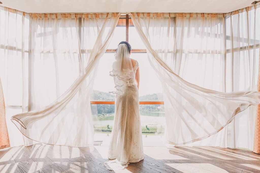 Bride in flowing gown gazes through sheer draped windows, showcasing Lake Garda wedding planner's attention to detail
