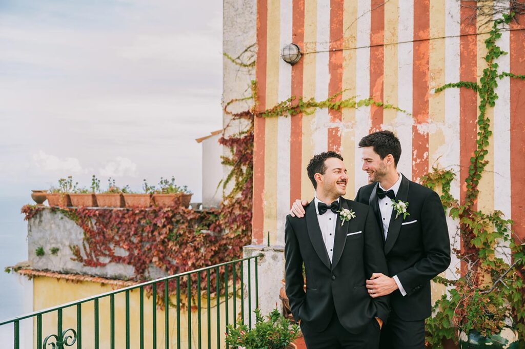 Two grooms in black tuxedos embrace affectionately outside Palazzo Confalone, Ravello, Italy