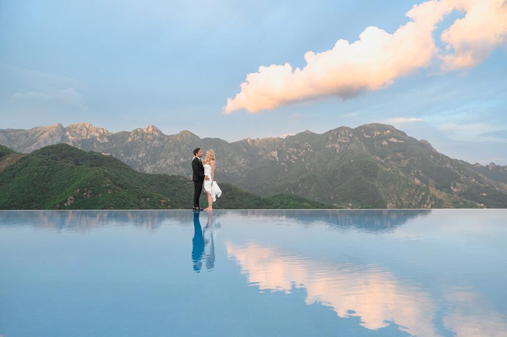 A Ravello wedding photographer captures newlyweds by an infinity pool, reflecting mountains at sunset