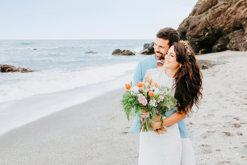 Italian Wedding Photographer shows a couple on a Taormina beach with a bouquet, capturing the coastal charm