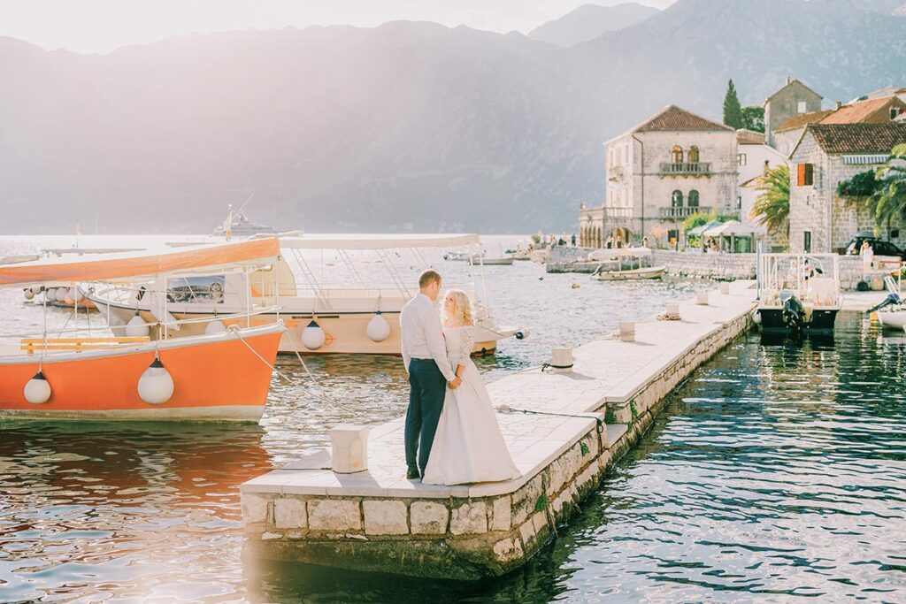 Italian Wedding Photographer captures a couple on a pier by an Italian lake, with a boat and lakeside town, showcasing the romantic scenery
